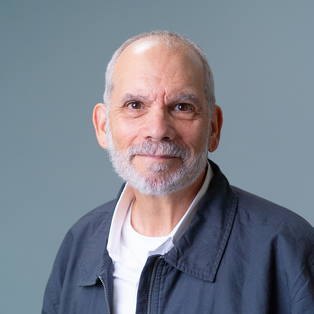 Professional headshot of a person with short gray hair and a gray beard, wearing a dark jacket over a white collared shirt, facing the camera against a soft gray-blue gradient background.