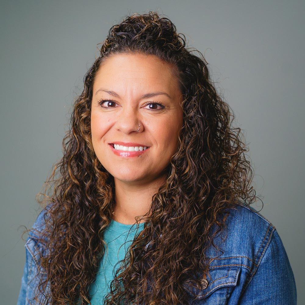 Professional headshot of a person with long curly hair, wearing a teal shirt and denim jacket, against a gray background.