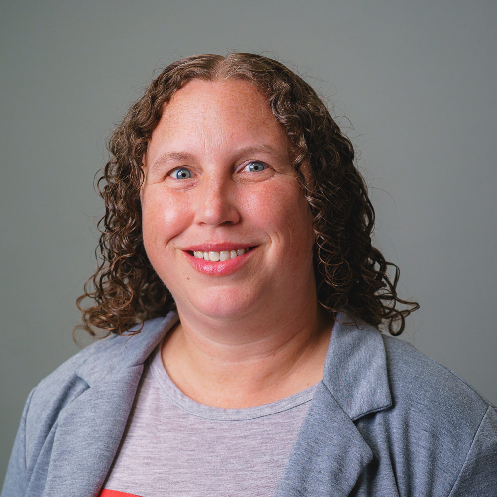 Professional headshot of a person with shoulder-length curly hair wearing a light gray blazer over a light-colored shirt, photographed against a light gray studio background. The image is framed from the chest up with soft, even lighting. Provide your feedback on BizChat Make it shorterUse plain, accessible languageDescribe the person’s expression.