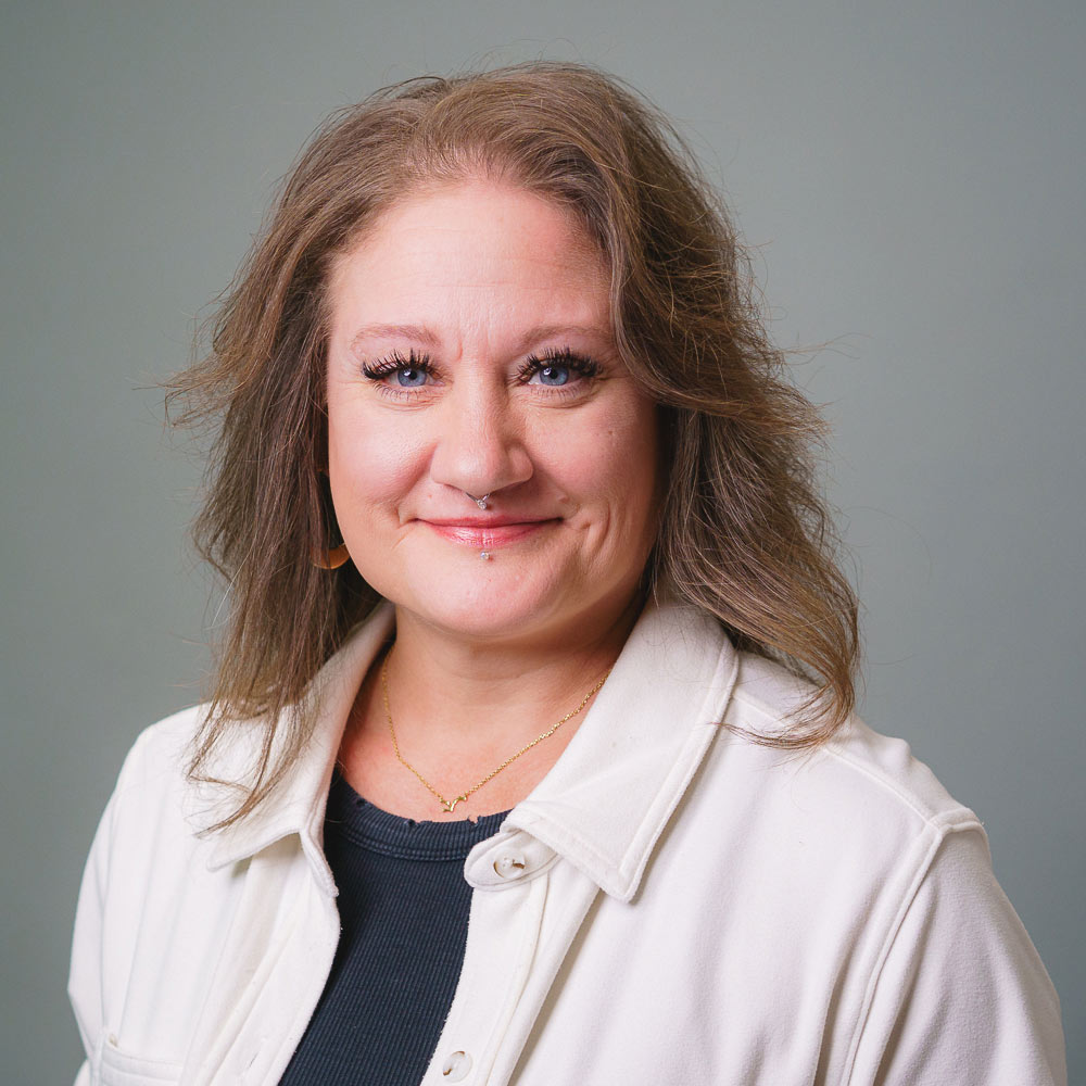 Professional headshot of a person with shoulder-length wavy hair, wearing a light-colored button-up overshirt over a dark top and a delicate necklace, photographed against a light gray studio background with soft, even lighting.