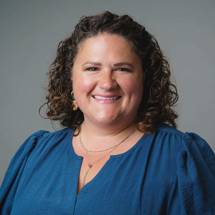 Professional headshot of a person with shoulder-length curly hair wearing a blue blouse, necklace, and colorful earrings, photographed against a plain gray background.