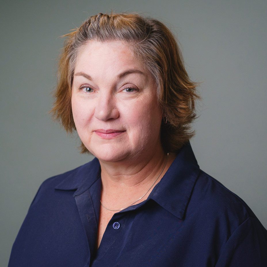 Professional headshot of a person with short hair wearing a navy blue collared shirt, photographed against a plain gray background.