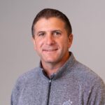 Professional headshot of a person wearing a gray quarter-zip pullover, photographed against a plain gray background.