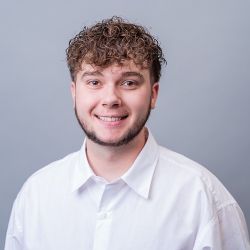 pagliarulo-tyler Professional headshot of a person with short curly hair wearing a white button-down shirt, facing the camera against a light gray background