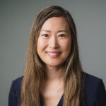 Professional headshot of a person with long hair wearing a dark blazer, photographed against a plain gray background.
