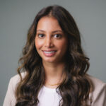 Professional headshot of a person with long wavy hair wearing a light-colored blazer and necklace, photographed against a plain gray background.