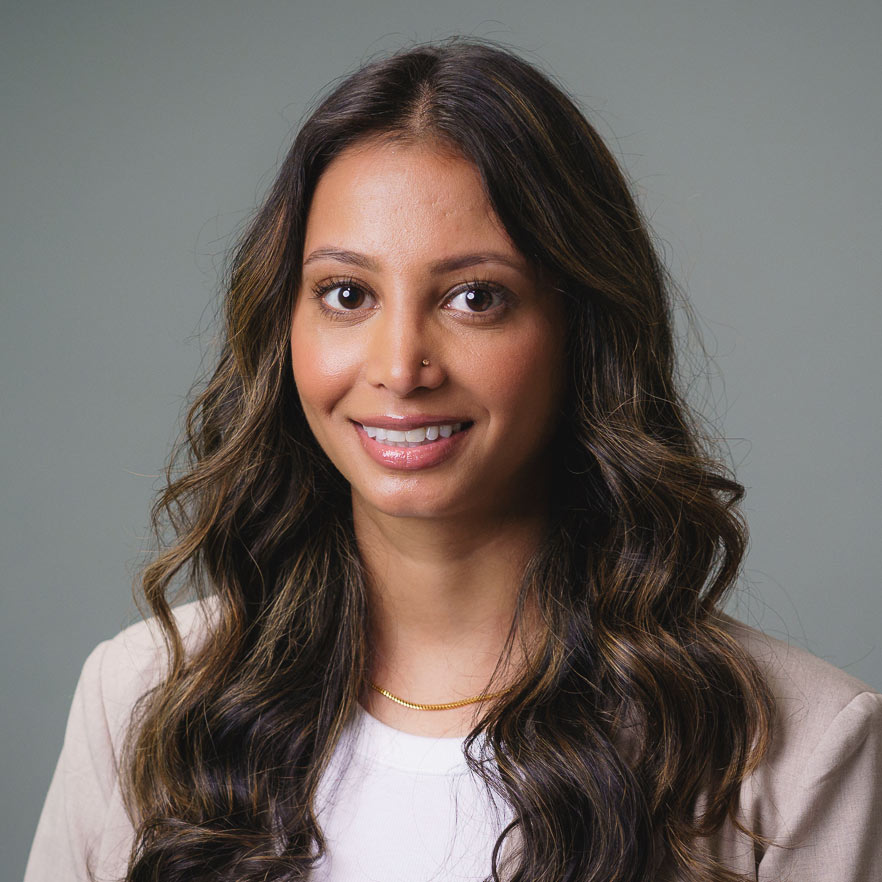 Professional headshot of a person with long wavy hair wearing a light-colored blazer and necklace, photographed against a plain gray background.