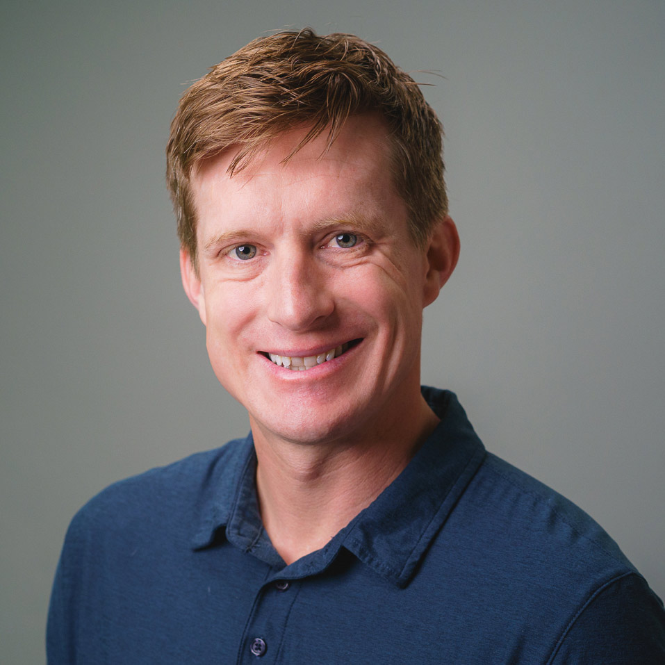 Professional headshot of a person with short light-brown hair wearing a blue collared shirt, photographed against a light gray studio background. The image is framed from the chest up with soft, even lighting.