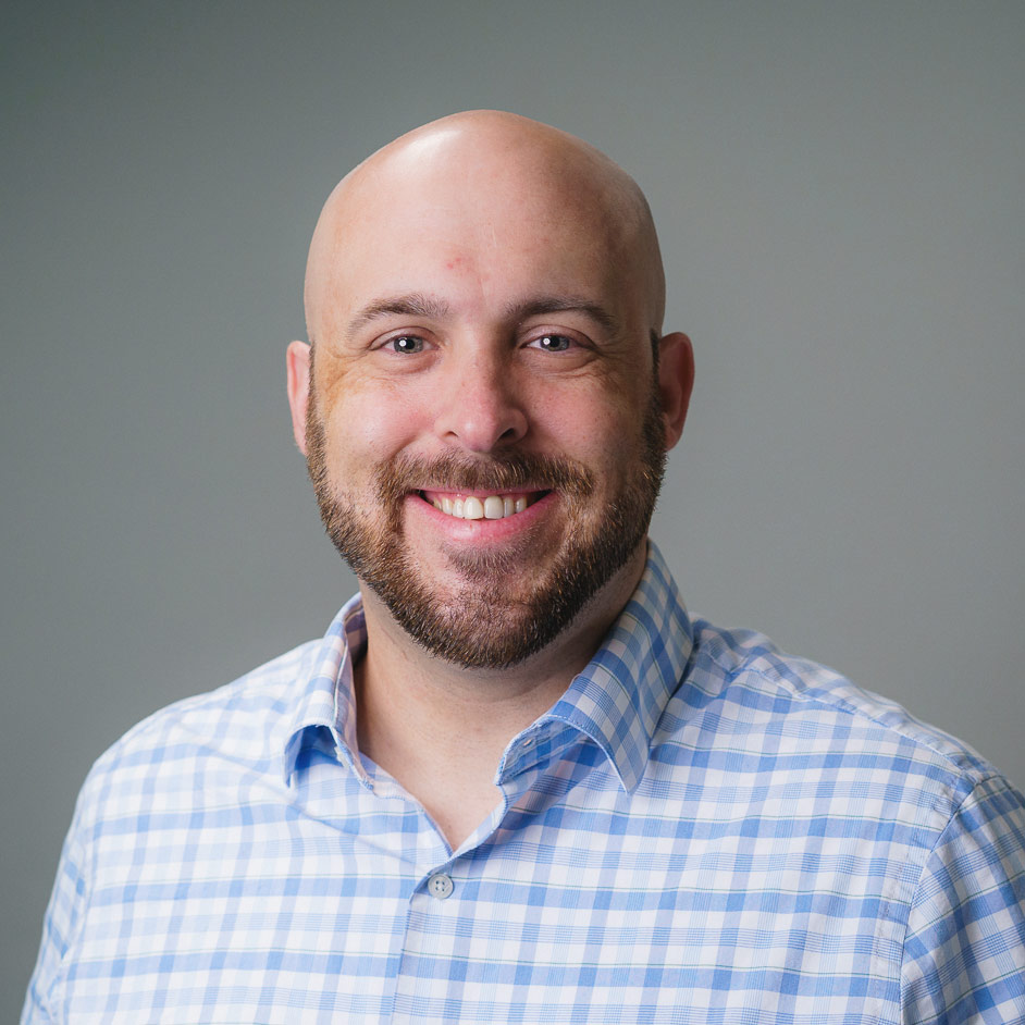 stanger-nate Professional headshot of a person with a shaved head and beard wearing a light blue checkered button-down shirt, photographed against a plain gray background.