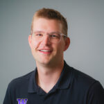 Professional headshot of a person wearing clear-framed glasses and a navy polo shirt with a purple logo, seated against a plain light gray background.