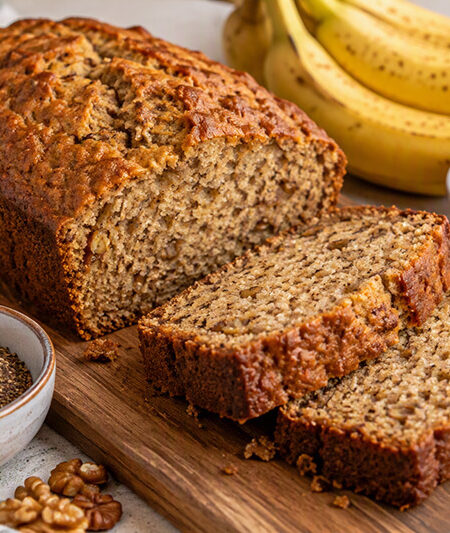 Freshly baked banana bread sliced on a wooden cutting board, surrounded by ripe bananas, rolled oats, flaxseed, and walnuts, showcasing a moist texture and golden-brown crust.