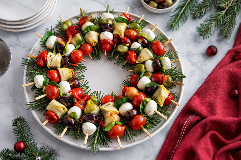Christmas wreath-shaped antipasto platter made with skewers of mozzarella, olives, cherry tomatoes, basil, artichoke hearts, and roasted peppers arranged on fresh greenery.