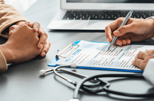 Close-up of a healthcare consultation with a patient seated across from a medical professional reviewing and signing paperwork on a clipboard. A stethoscope and laptop are visible on the desk.