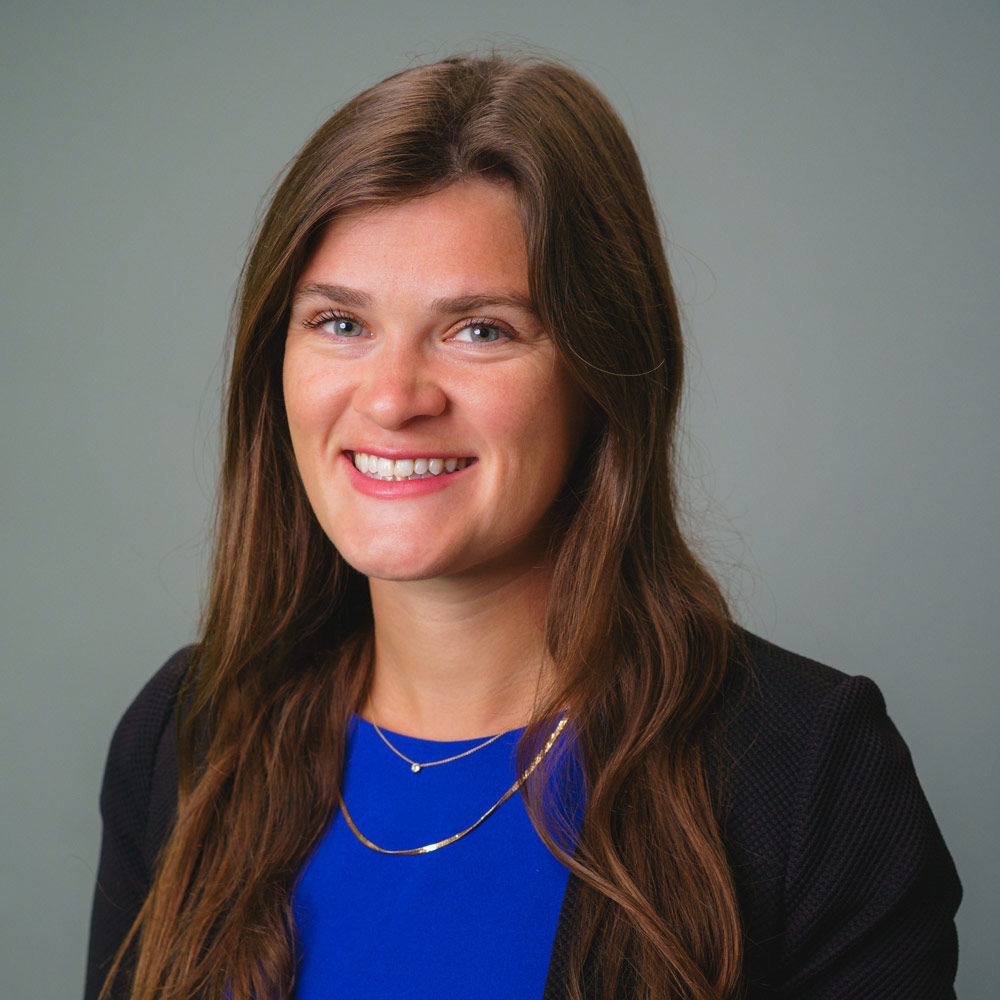 Headshot of a person with long brown hair wearing a blue top and dark blazer against a gray background.