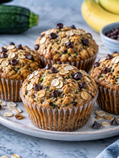Zucchini muffins topped with oats and chocolate chips on a gray plate with fresh zucchini and oats nearby.