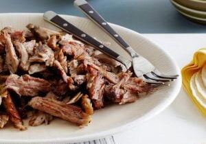 White plate filled with shredded pork carnitas with crispy, browned edges, two metal forks resting on the plate, placed on a white surface beside a folded yellow napkin.