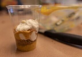 Pumpkin pie parfait served in a clear plastic cup, layered with pumpkin filling, whipped cream, and graham cracker crumbs on a kitchen counter.
