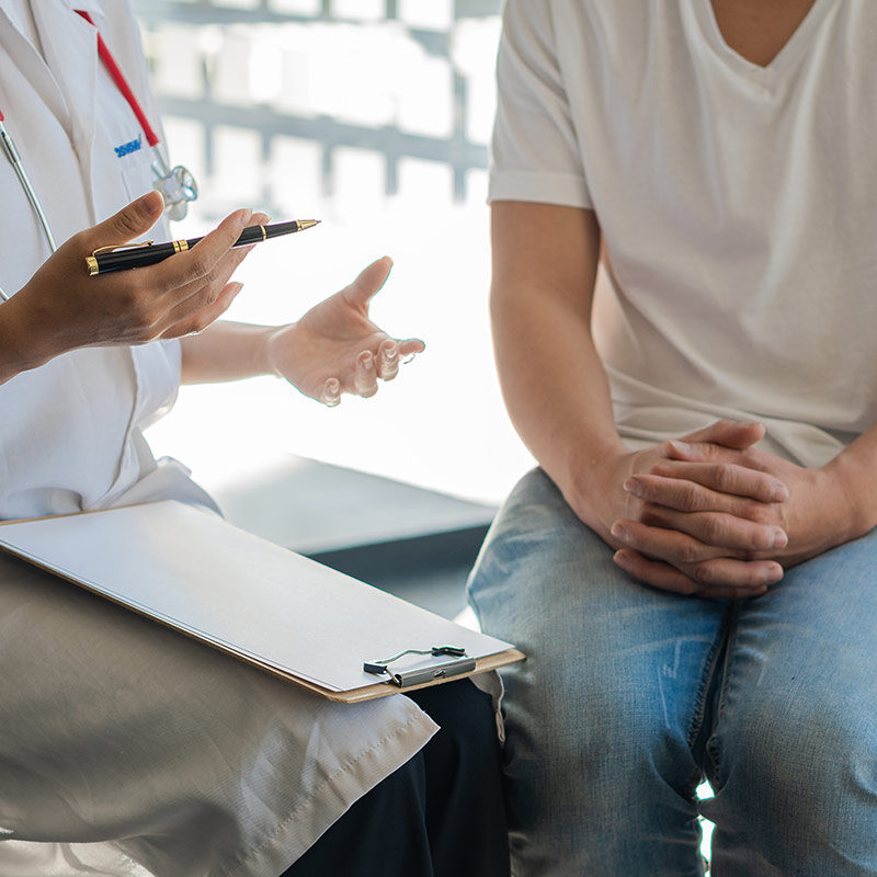 Healthcare provider with a stethoscope and clipboard speaking with a patient seated in an exam room during a medical consultation. The provider gestures while holding a pen, and the patient sits with hands clasped.
